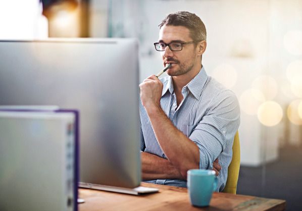 man staring at computer screens thinbking with pen in mouth